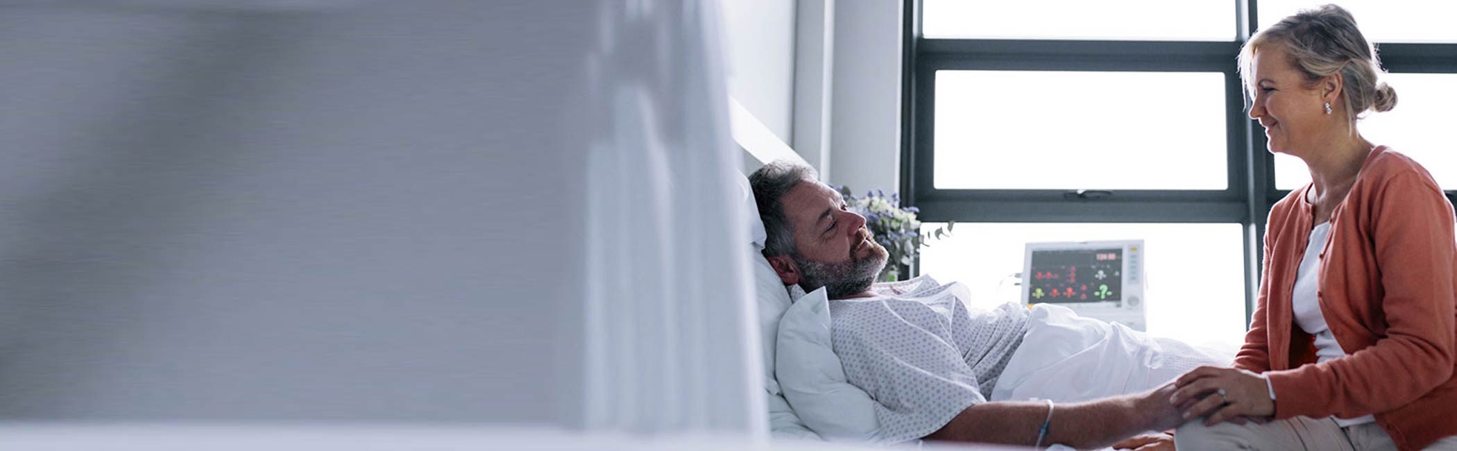 Kvinde sidder og holder sin sengelagte mand i hånden på et hospital.