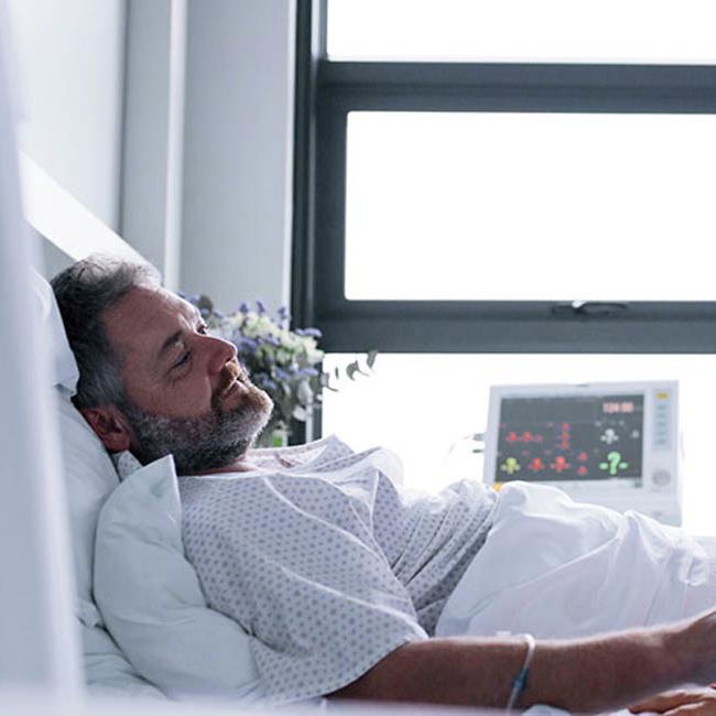 Kvinde sidder og holder sin sengelagte mand i hånden på et hospital.