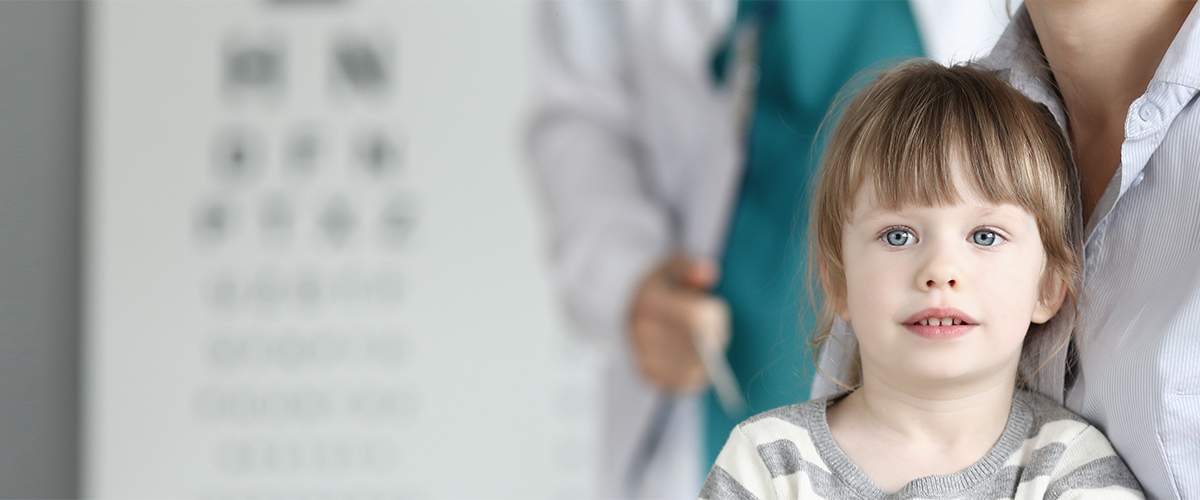 Barn kigger ind i kameraet. I baggrunden ses mor og børnelæge.