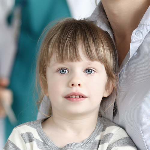 Barn kigger ind i kameraet. I baggrunden ses mor og børnelæge.