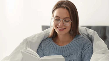 Glad ung kvinde sidder i en seng med dynen om sig. Hun kigger ned på noget.