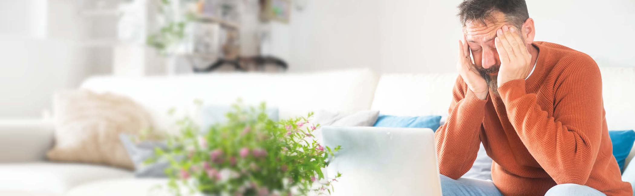 Træt mand sidder i en sofa med fingrene i tindingerne og ser ned på sin computer.