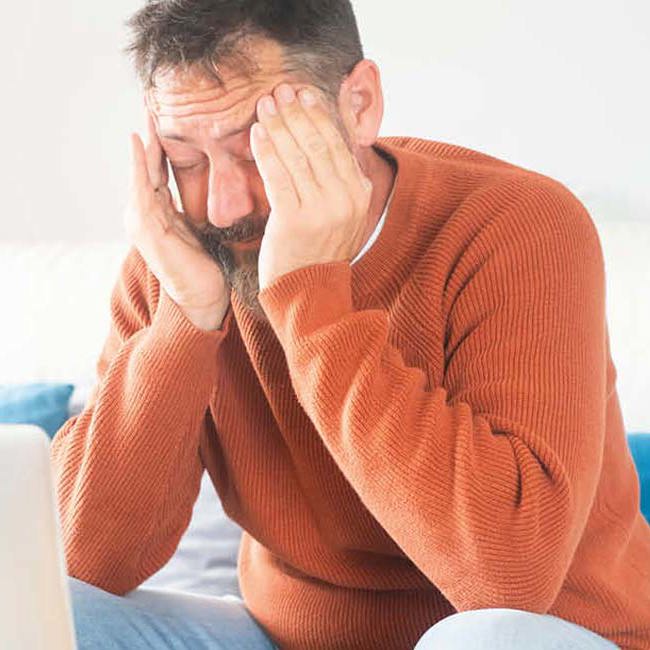 Træt mand sidder i en sofa med fingrene i tindingerne og ser ned på sin computer.