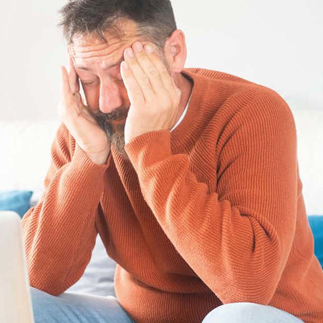 Træt mand sidder i en sofa med fingrene i tindingerne og ser ned på sin computer.