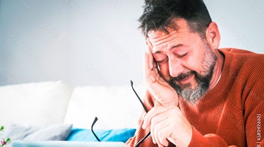 Træt mand sidder i en sofa med fingrene i tindingerne og ser ned på sin computer.