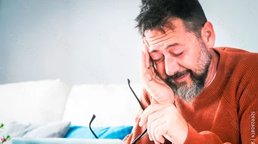 Træt mand sidder i en sofa med fingrene i tindingerne og ser ned på sin computer.