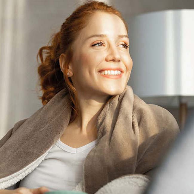 Glad kvinde sidder i en sofa med et tæppe om sig og smiler.