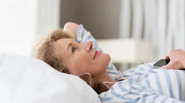 Glad kvinde ligger på ryggen i sengen med høretelefoner på.