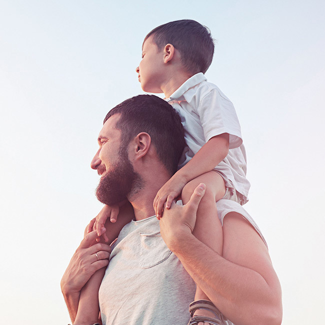 Far står med sin søn på skuldrene ude i naturen.